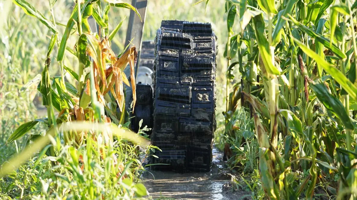 A head-on perspective looking straight at a muddy airless EWRS-Pivot-XT tire as it moves through tall, wet grass and a cornfield row. The high-traction tread design is front-and-center, highlighting its ability to pull through anything